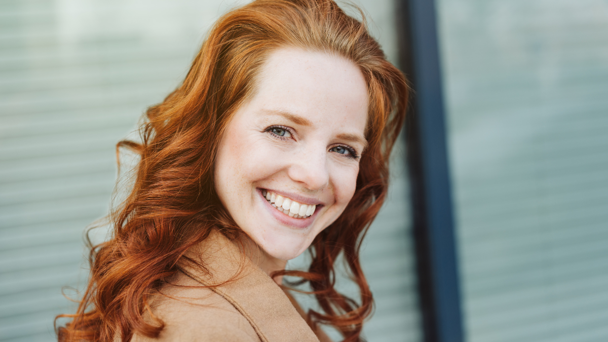 Smiling pretty woman with curly red hair close-up half-turn portrait. Gorgeous lady in beige coat and with beautiful smile. Glass wall or window blurred in background.