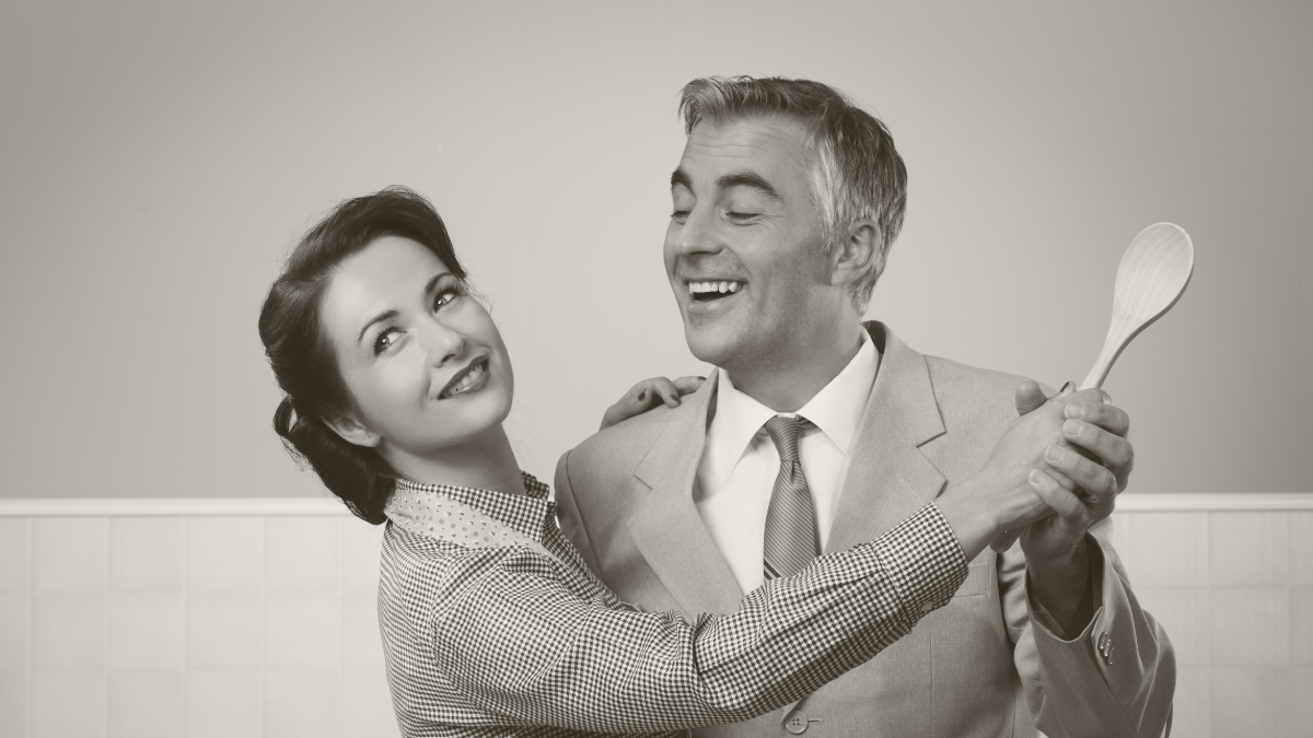 Smiling vintage couple dancing in the kitchen and holding a wooden spoon.