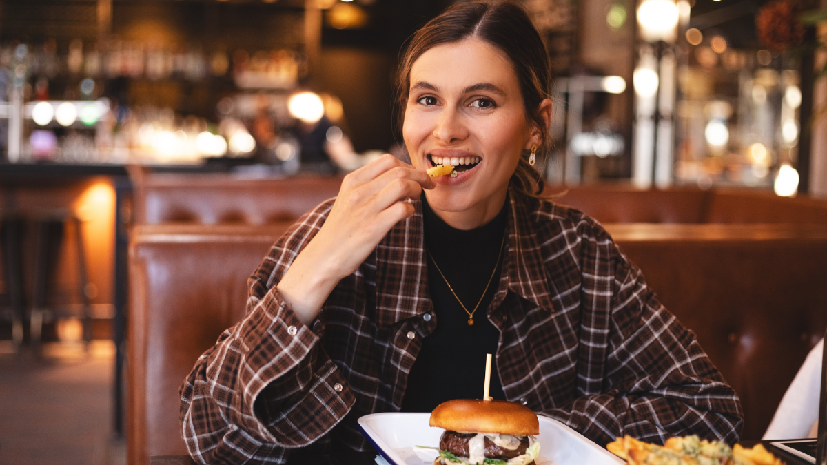 Fast food lover. Happy lady eating burger and french fries, biting potatoes and looking camera, sitting in restaurant. Playful woman holding french fries, burger in plate and tasty. Cheat meals.