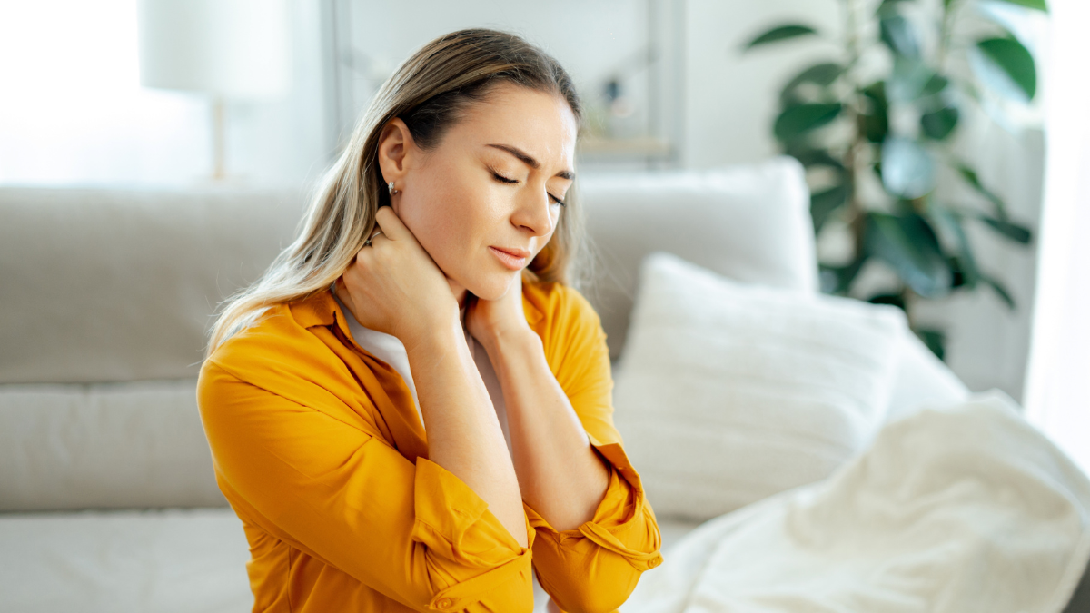 Concept of neck pain, stiffness, or muscle tension. Caucasian exhausted woman in a yellow shirt sitting on a cozy couch at home, holding her neck with a pained facial expression.