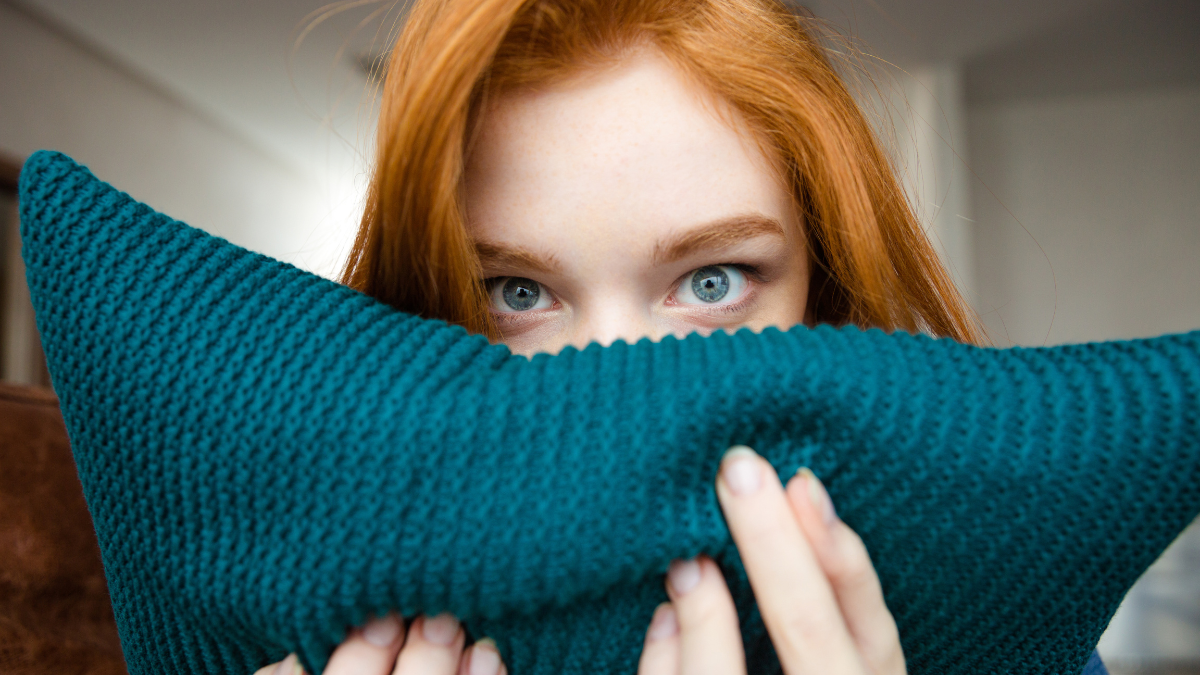 Shy pretty young woman with red hair hiding face behind knitted pillow.