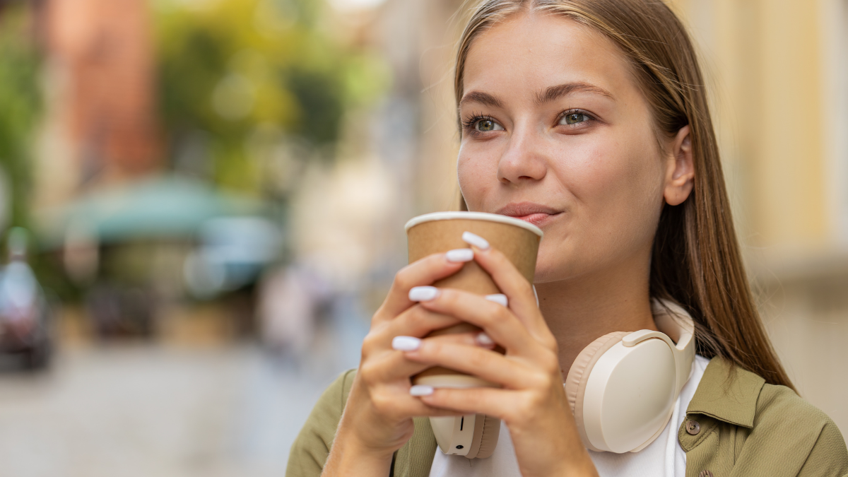 Happy Caucasian woman enjoying morning coffee hot drink and smiling. Relaxing, taking a break. Young girl teenager walking on urban city center street, drinking coffee to go. Town lifestyles outside.