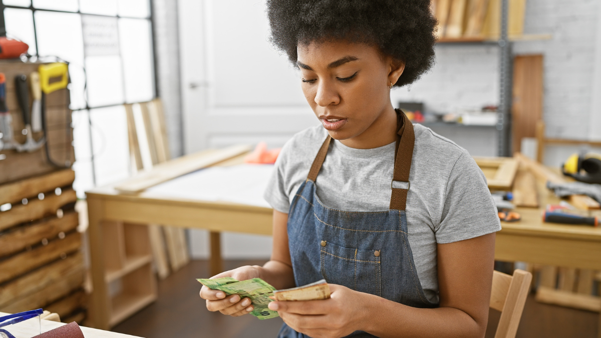 A black woman counts south african rand in a woodworking workshop, reflecting a business or financial theme.