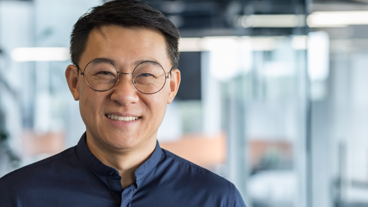 Close up photo portrait of successful young Asian investor, man in glasses smiling and looking at camera in shirt, businessman working inside modern office building, satisfied accountant financier.