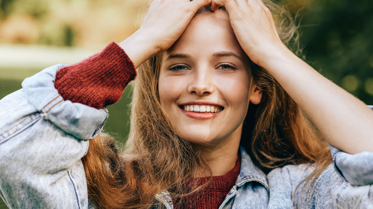 Close-up portrait of beautiful happy young woman smiling broadly with healthy toothy smile, wearing red sweater and denim jacket, posing on nature background in the park. People, travel, lifestyle.