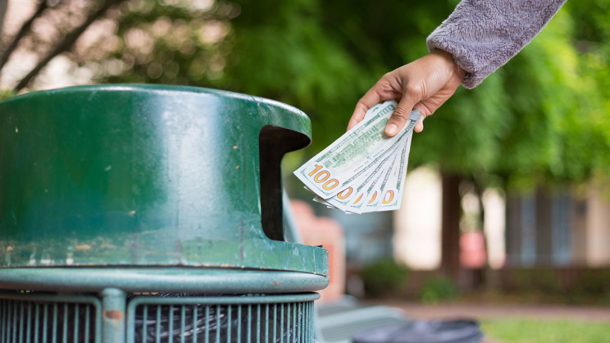 Closeup cropped portrait of someone hand tossing cash dollar bills money, hundred dollar bills in trash can, isolated outdoors green trees background. Bad financial investment decisions concept.
