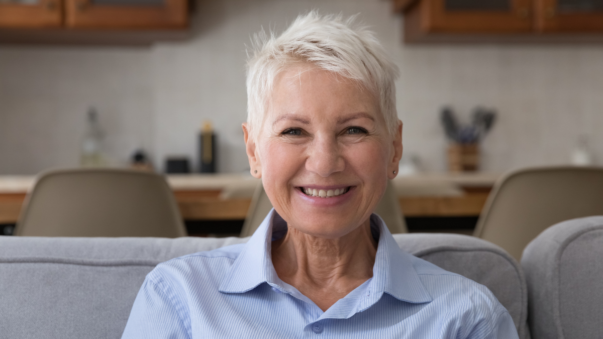 Head shot portrait older blonde woman resting on sofa staring at camera feels happy, having wide toothy smile and attractive appearance, enjoy retired carefree life at home alone looking optimistic.