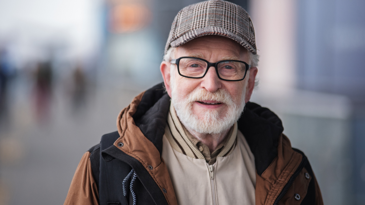 Old generation. Portrait of positive senior bearded man with glasses is standing outdoors and looking at camera with slight smile.