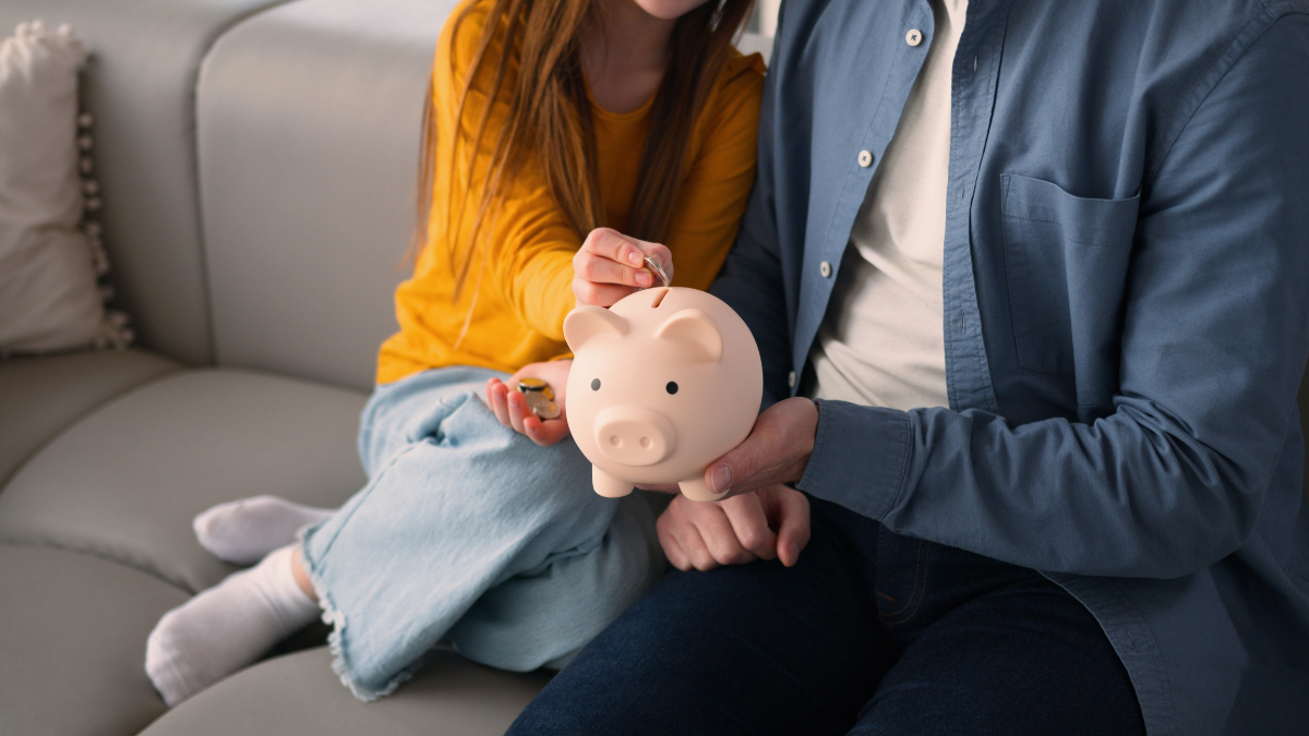 Happy Caucasian family little schoolgirl child girl daughter kid saving money at home guy parent dad male father man put coins in piggy bank savings economy financial education invest charity donation.