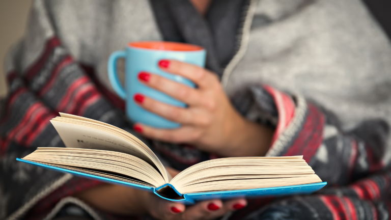 Woman reading a book and holding a mug of hot beverage.