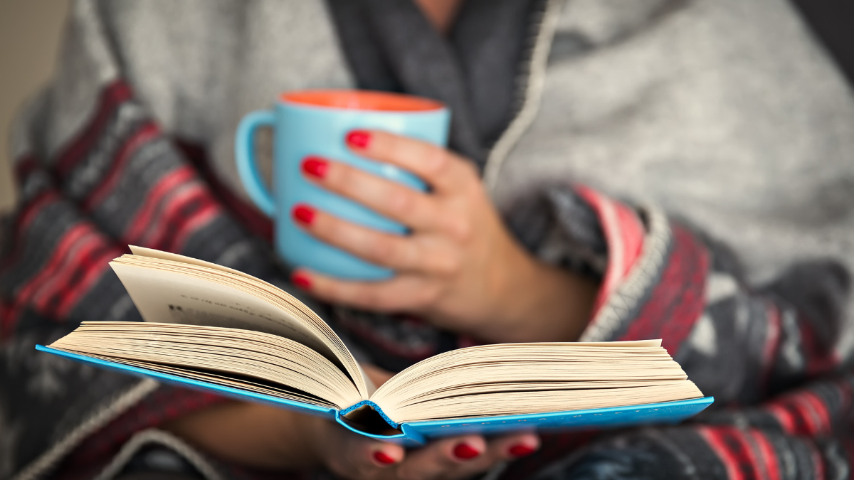 Woman reading a book and holding a mug of hot beverage.
