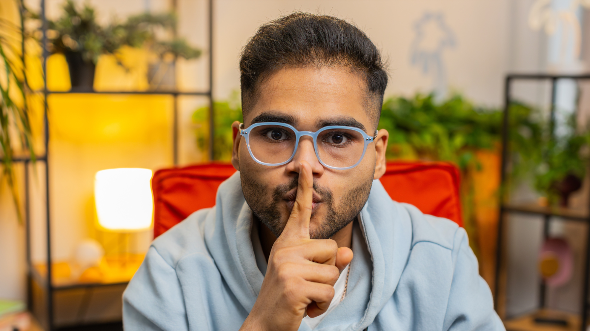 Shh be quiet please. Indian young man presses index finger to lips makes silence hush gesture sign do not tells gossip secret. Bearded Arabian guy at home apartment living room sitting on chair.