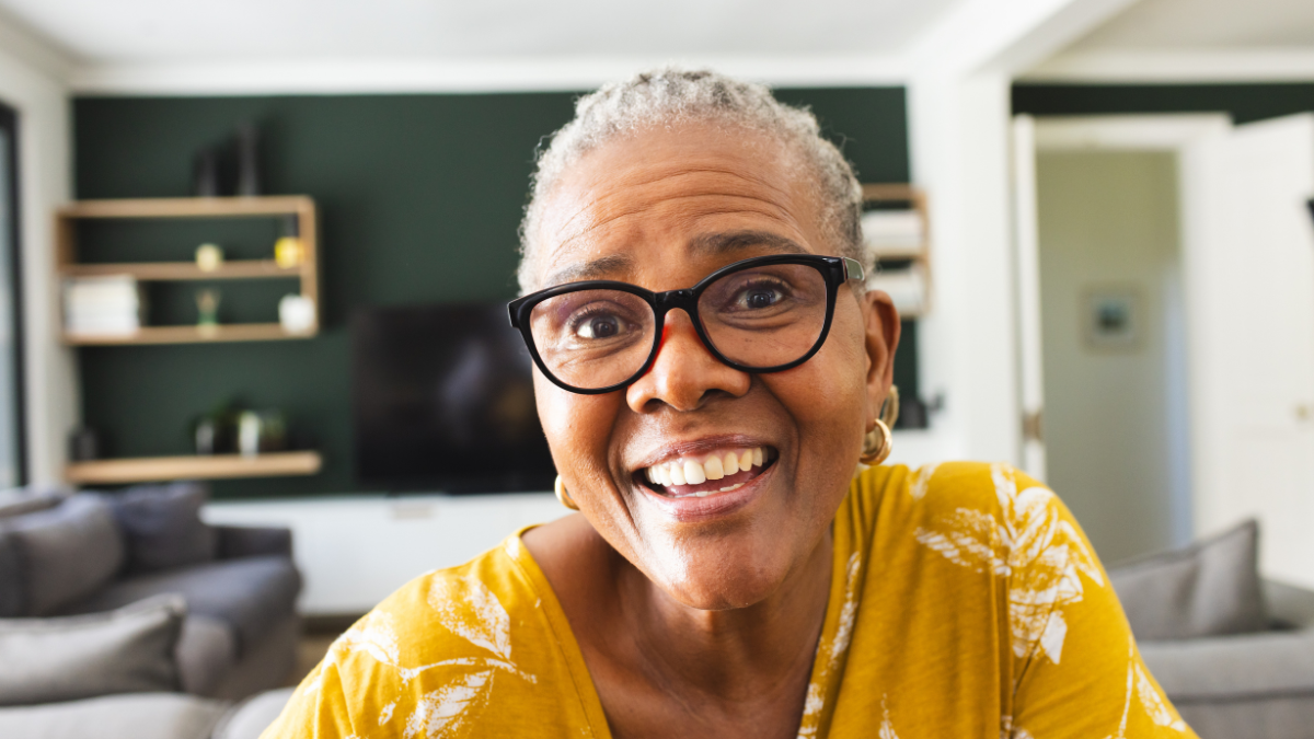 Senior African American woman smiles warmly on video call, wearing glasses and a yellow top. Her joyous expression brightens the cozy home setting.