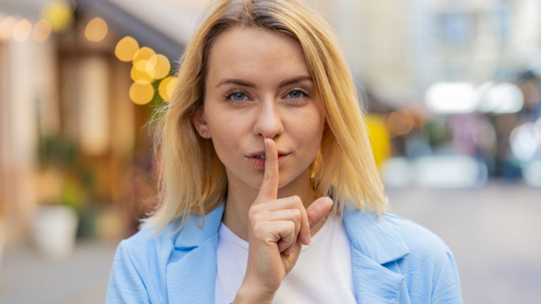 Shh be quiet please. Blonde young woman presses index finger to lips makes silence hush gesture sign do not tells gossip secret outdoors. Young lady female stand on urban city street. Town lifestyles.