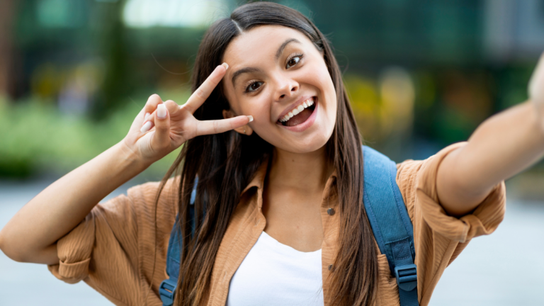 Cute pretty long-haired brunette young lady zoomer taking selfie outdoors, gesturing peace and smiling, walking by street, going to university, college, posting on social media, copy space.
