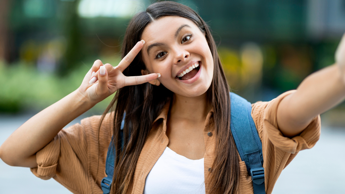 Cute pretty long-haired brunette young lady zoomer taking selfie outdoors, gesturing peace and smiling, walking by street, going to university, college, posting on social media, copy space.