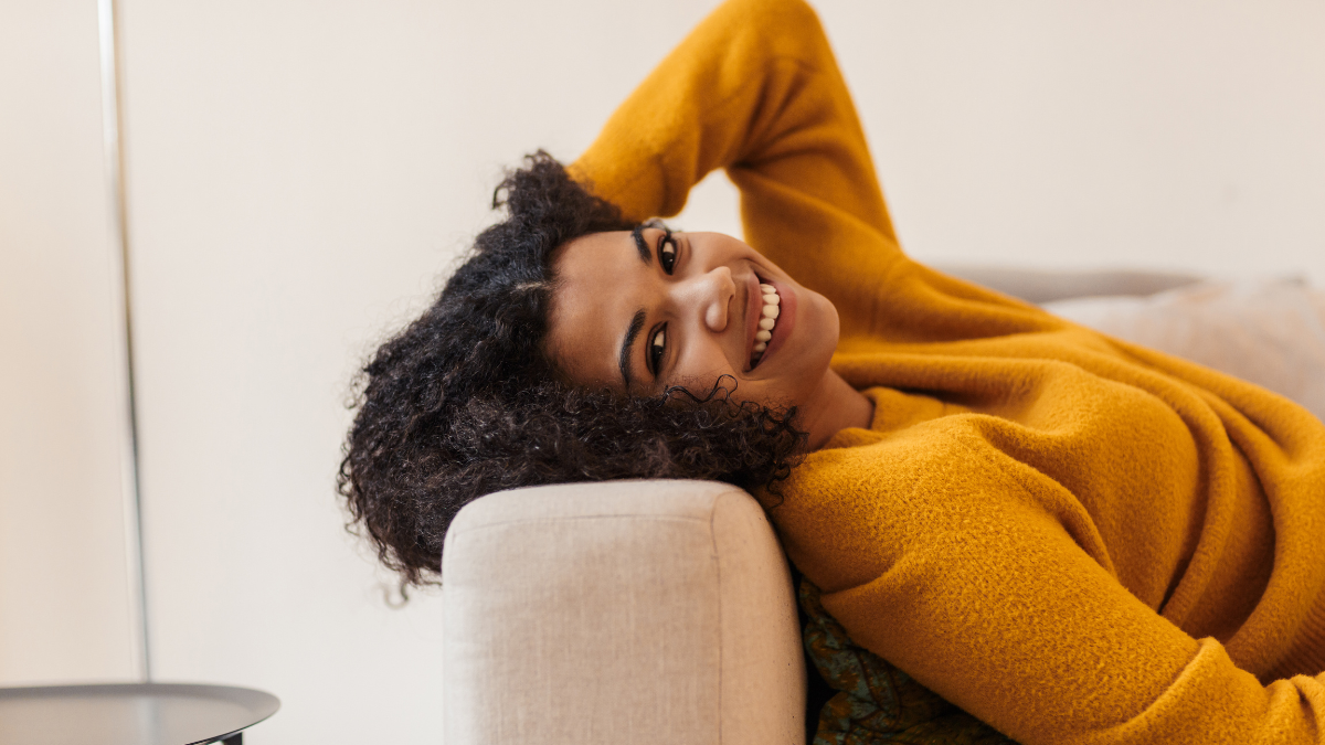 Image of joyful nice african american woman smiling while lying on couch in living room.