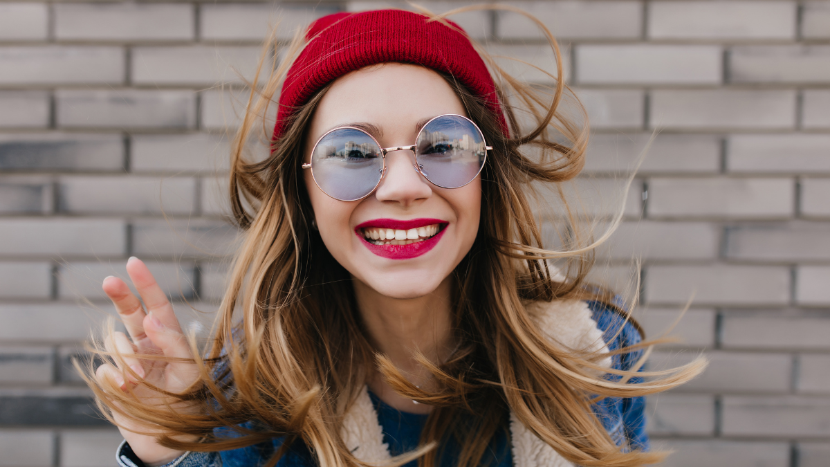 Lovable woman in casual blue glasses fooling around during outdoor photoshoot. Portrait of excited blonde girl with bright makeup posing on brick background.