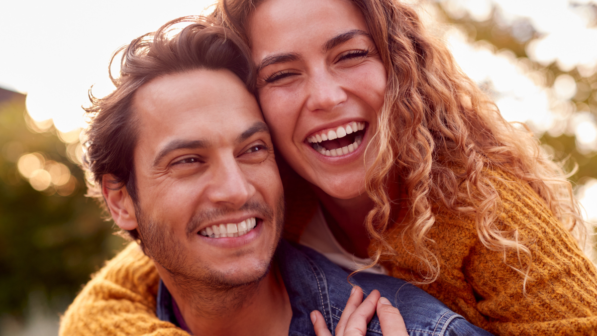 Portrait Of Happy Loving Couple With Man Giving Woman Piggyback As They Hug In Autumn Park Together.