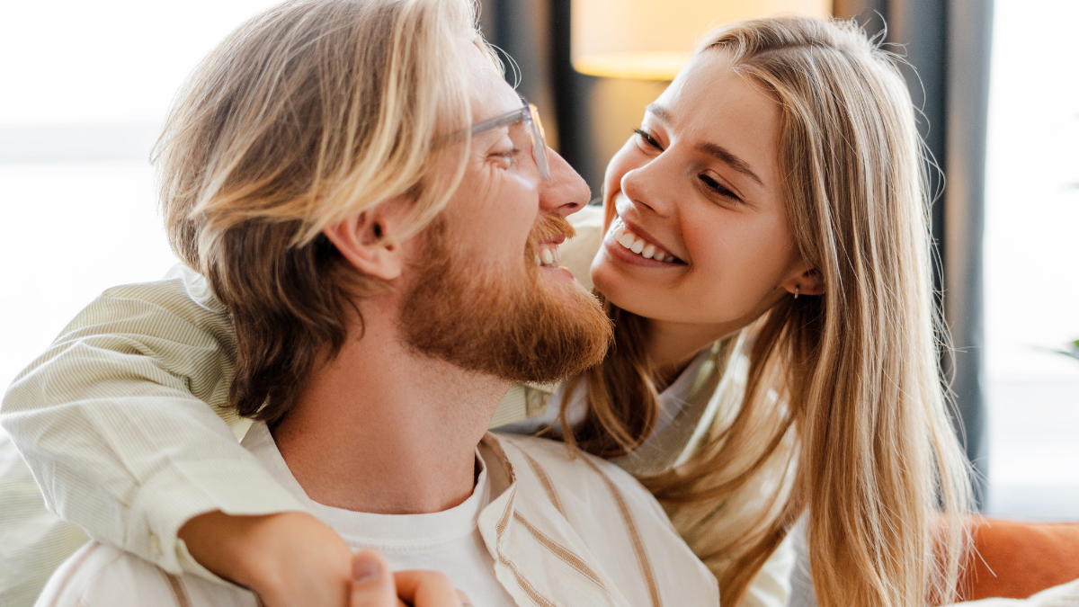 Happy young couple cuddling on the sofa at home, smiling and showing love. Blond woman and bearded man in cozy living room, enjoying each other's company. Joyful and romantic moment captured.