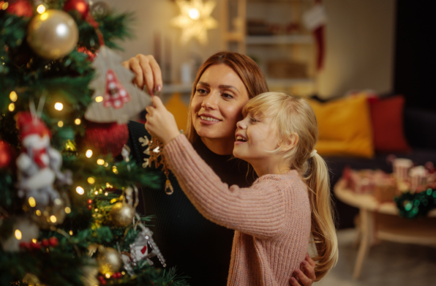 Happy Caucasian mother and her daughter are in their home during Christmas, decorating a Christmas tree and having fun.