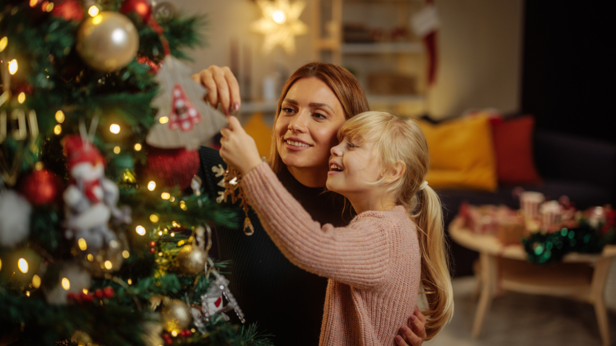 Happy Caucasian mother and her daughter are in their home during Christmas, decorating a Christmas tree and having fun.