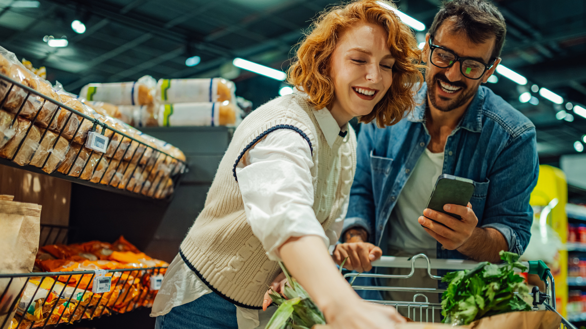 Young couple shopping groceries together in supermarket, using smartphone and choosing products, putting groceries in shopping cart, smiling and having fun.