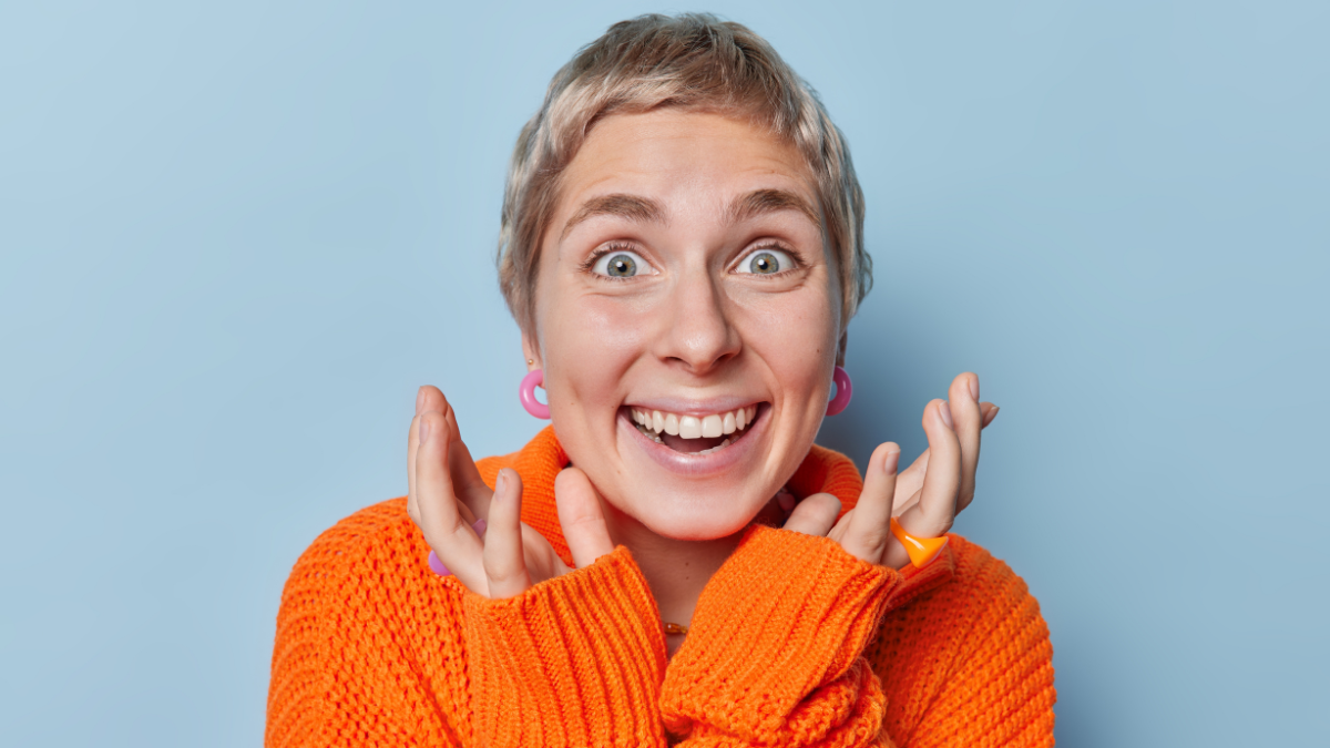Beautiful short haired European woman reacts to something amazing keeps hands near face smiles broadly shows perfect white teeth dressed in loose knitted jumper isolated over blue background.