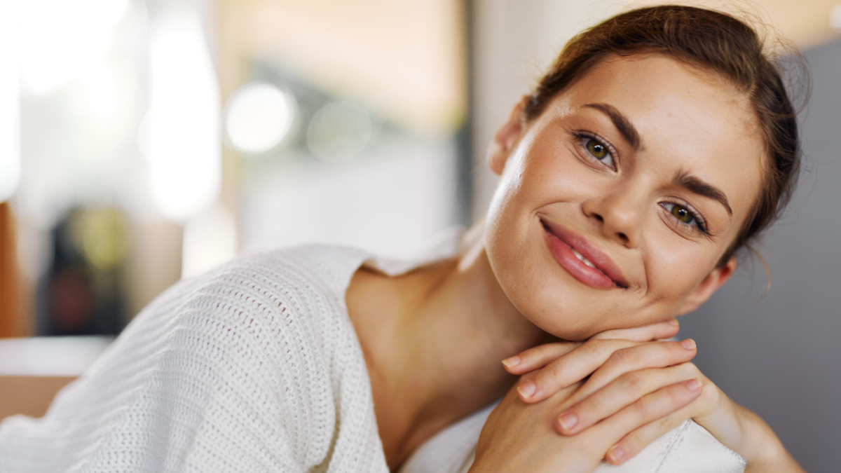 Happy Woman Sitting on a Cozy Couch at Home, Relaxing and Smiling A Beautiful Young Brunette with a Joyful Smile, Wearing a Comfortable Sweater, Resting in a Warm and Attractive Living Room She Looks because she has frugal living tips that won't leave you feeling deprived.