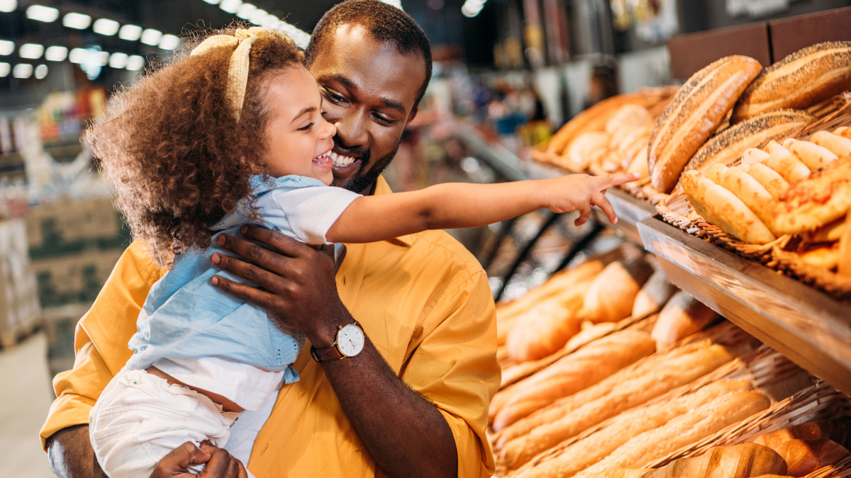 african american little child pointing by finger at pastry to father in supermarket while they save money using grocery savings apps.