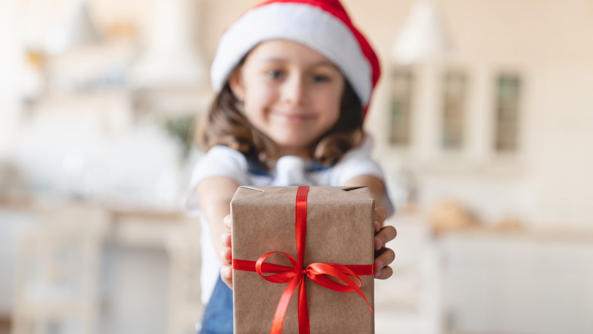 Focused close up shot of little Santa`s helper. Caucasian small kid child girl holding Christmas present gift box looking at camera, celebrating Christmas New Year at home.