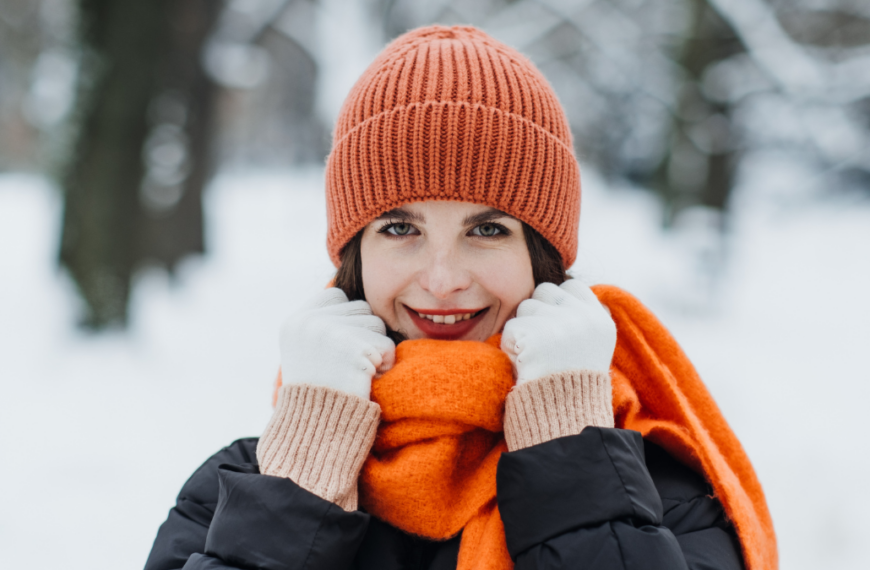 Woman in orange winter hat and scarf smiling in snow-covered park. Warm seasonal fashion, winter style trends, cozy outdoor clothing. Color Therapy in Seasonal Fashion.