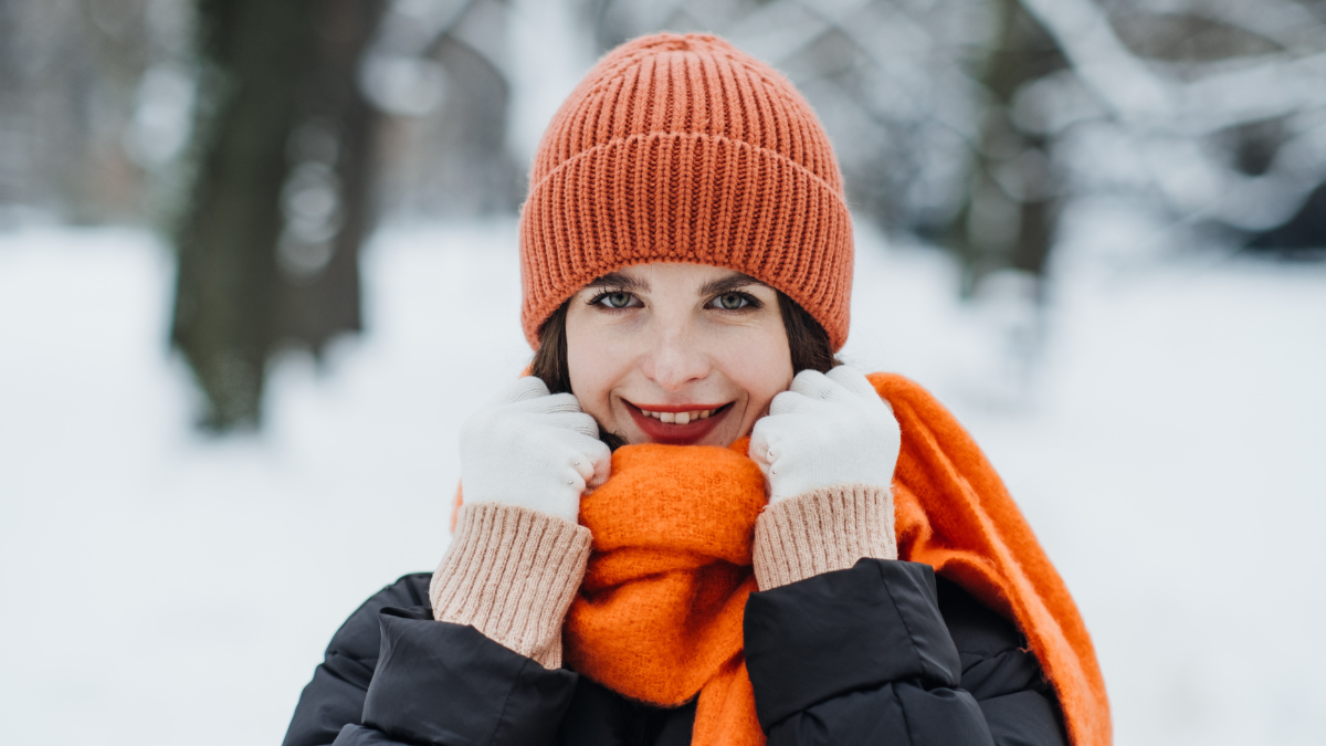 Woman in orange winter hat and scarf smiling in snow-covered park. Warm seasonal fashion, winter style trends, cozy outdoor clothing. Color Therapy in Seasonal Fashion.