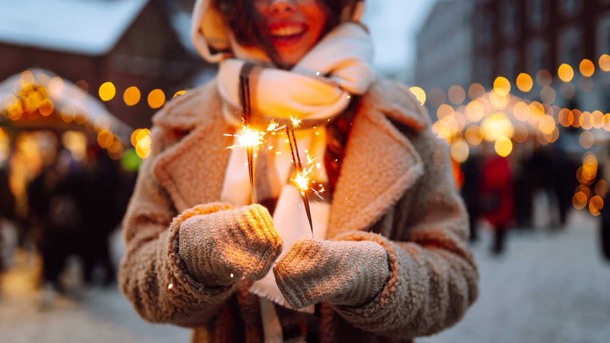 Close up Christmas sparkles in hands. Young smiling woman holding burning sparkler in cold winter evening on street Christmas market. Christmas, new year.
