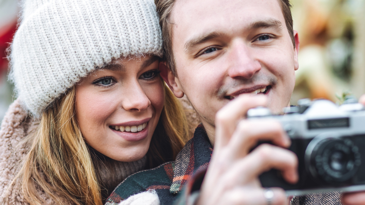 Beautiful young loving couple, boyfriend and girlfriend having fun on a Christmas market wearing warm clothes, taking pictures on old retro camera. Outdoors, winter time, snowy weather.