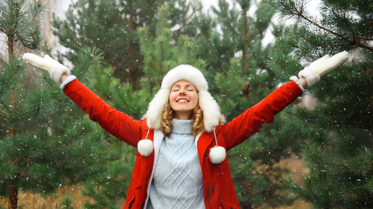 Winter day, happy young woman enjoys snow, warm weather against Christmas tree, joyful girl in hat, red jacket raising her hands up in snowy green forest with snowflakes.