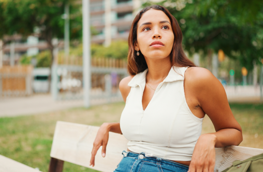 Lovely tanned woman with long brown hair wearing white top sitting relaxed on park bench looking away. Life style concept. Slow motion.