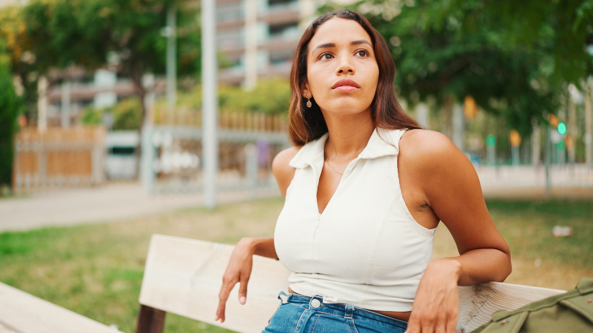 Lovely tanned woman with long brown hair wearing white top sitting relaxed on park bench looking away. Life style concept. Slow motion.