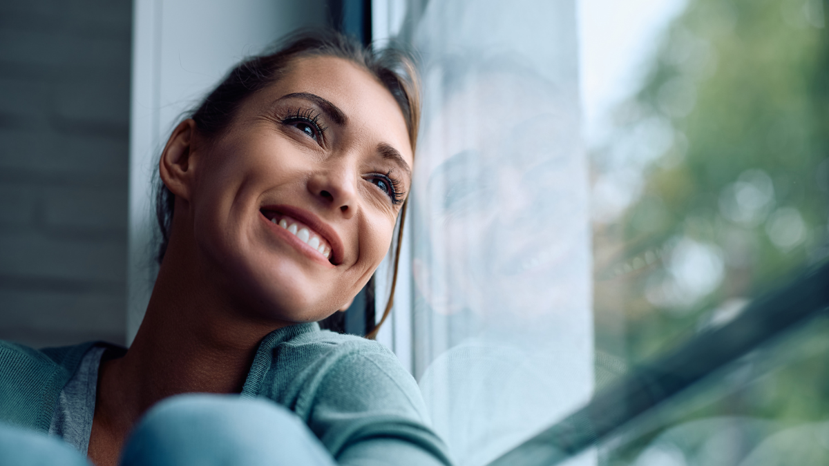 Smiling woman contemplating while sitting by the window at home and looking through it.