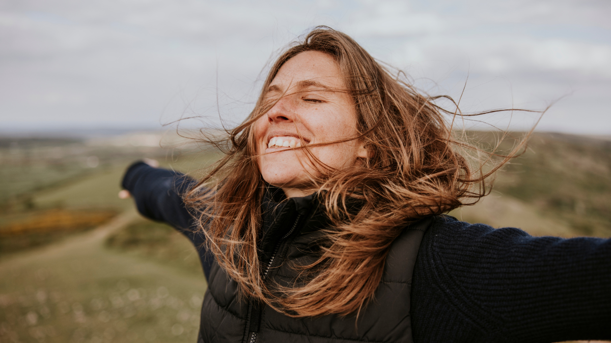 A happy blond woman with brown hair, arms outstretched, enjoying the nature outdoors. The happy woman is smiling, embracing the wind, and feeling free in nature. Happy woman outdoors in nature.