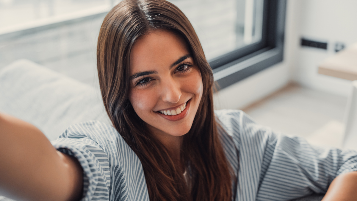 Happy attractive young adult woman housewife sitting on sofa looking at camera in modern cozy home, smiling single 20s lady beautiful face taking a selfie alone enjoy wellbeing posing for portrait.