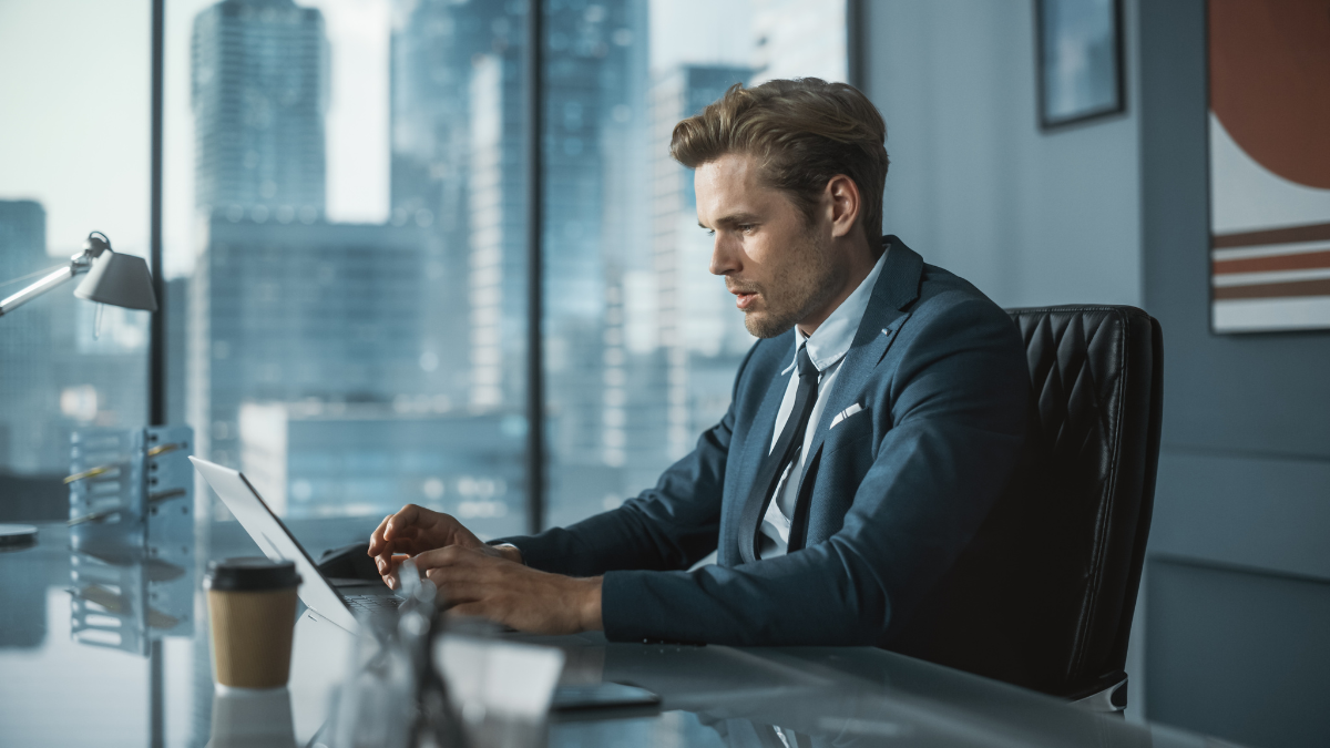 Confident Businessman in a Suit Sitting at a Desk in Modern Office, Using Laptop Computer, Next to Window with Big City with Skyscrapers View. Successful Finance Manager Planning Work Projects..