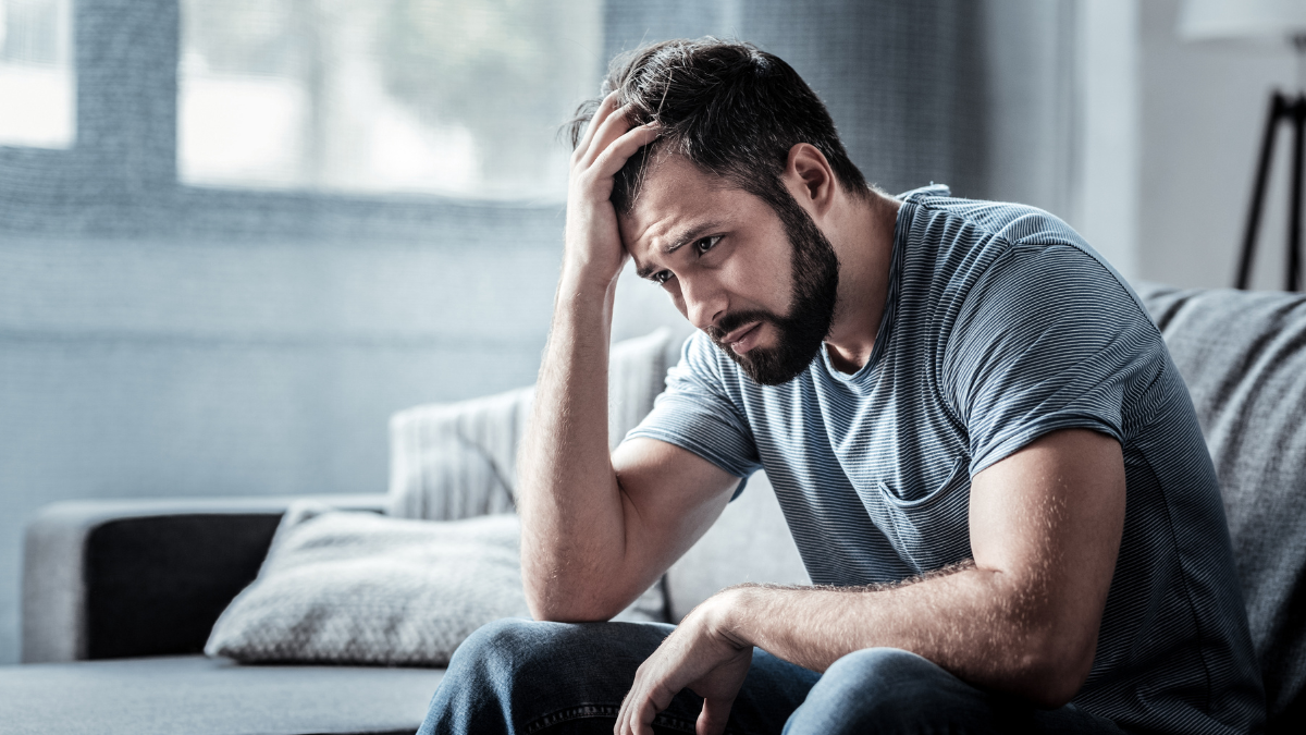 Unpleasant pain. Sad unhappy handsome man sitting on the sofa and holding his forehead while having headache.