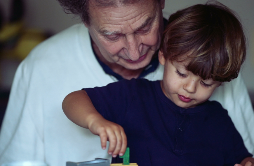 Candid grandson and grandfather bonding moment child plays with with car toy on grandparent lap.