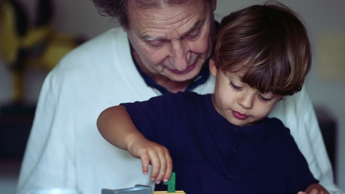 Candid grandson and grandfather bonding moment child plays with with car toy on grandparent lap.