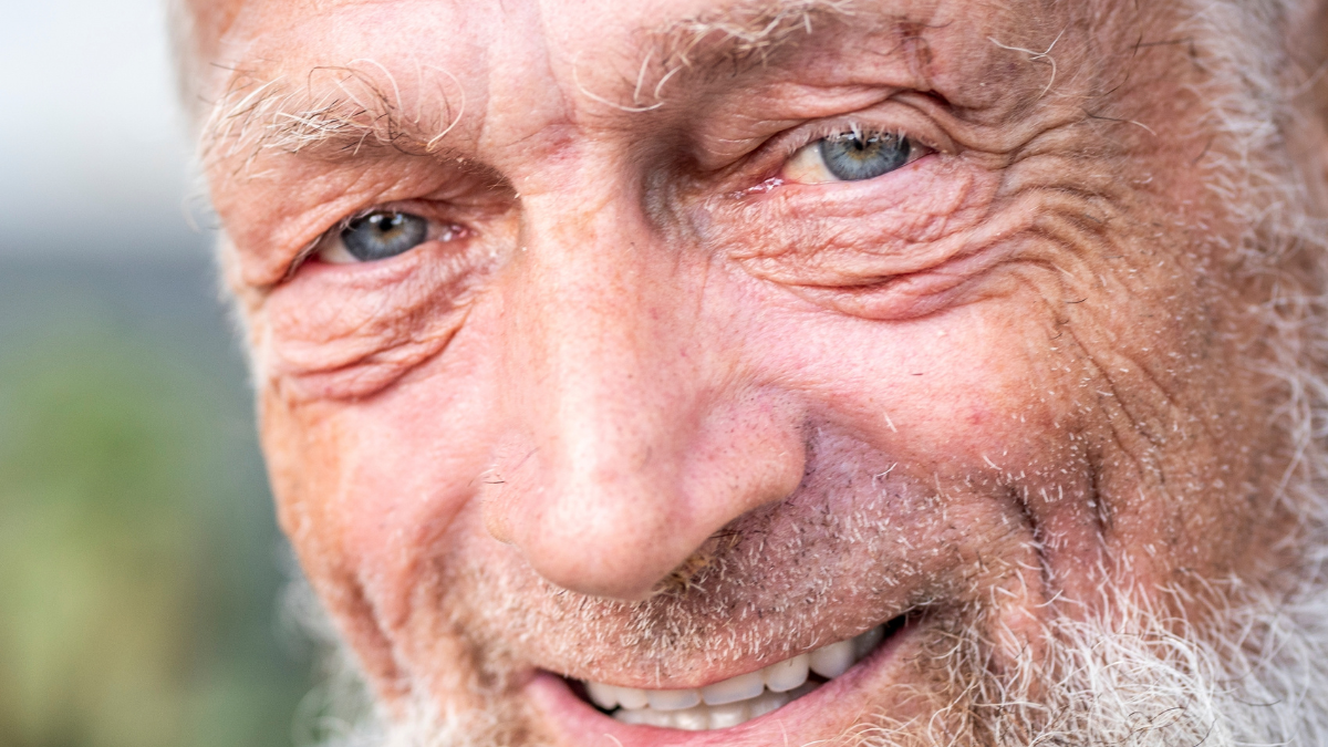 Close up portrait of happy 70-year-old optimist man with smiling wrinkled face, a touch of sadness feeling in his blue eyes. Park outdoors background defocused. Elderly people smile at the camera.