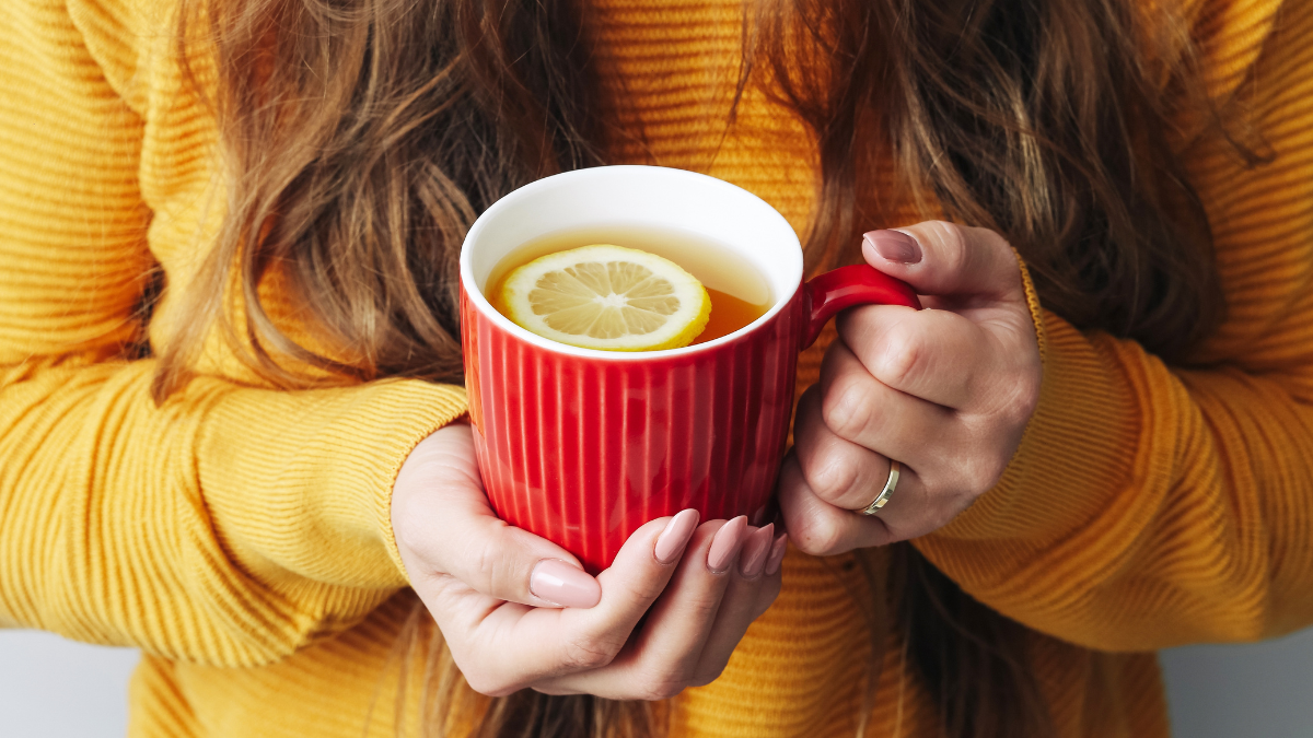 Hot tea in red mug. Woman holding cup of lemon tea. Cold winter background. Yellow woolen sweater warm clothing. Lemon slice in a tea mug. Female hands holding tea. Ceramic mug in hands.