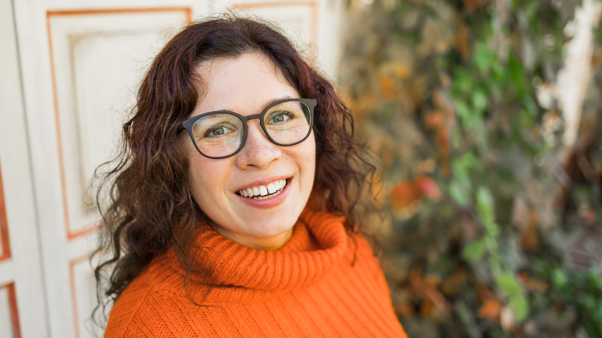 Happy portrait of attractive young woman in stylish glasses in knitted fashionable orange sweater on background of leaves copy space and empty space for text. Girl walks in city.