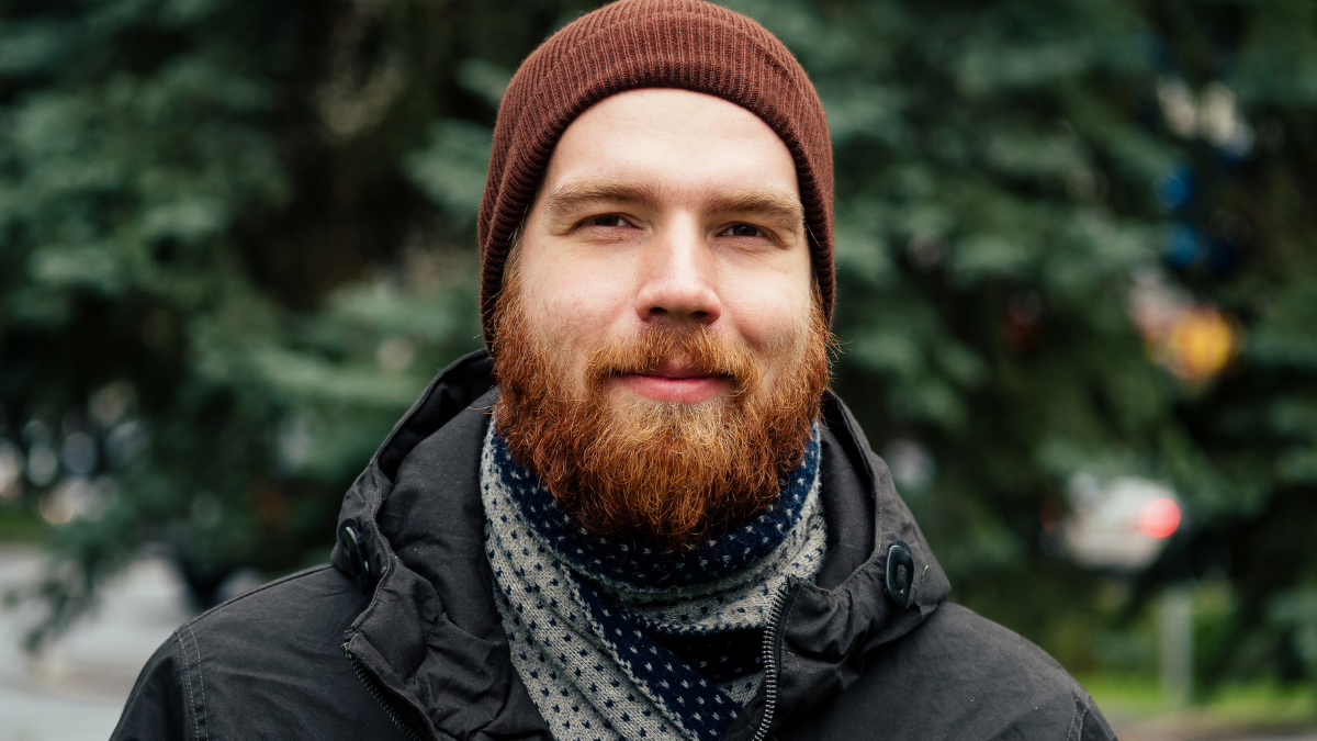 man with a red beard and hat on a background of green trees.