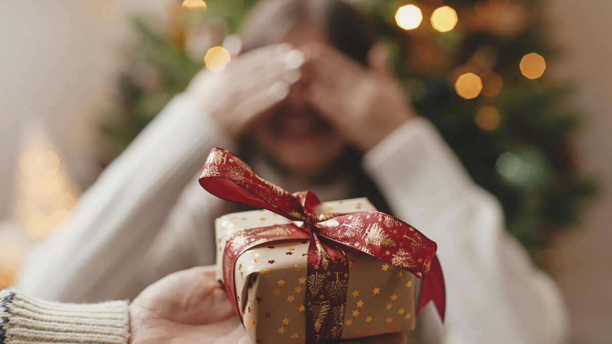 Merry Christmas and Happy Holidays! Hand holding christmas gift box on background of happy woman closing eyes waiting for surprise. Exchanging presents at christmas tree in atmospheric room.